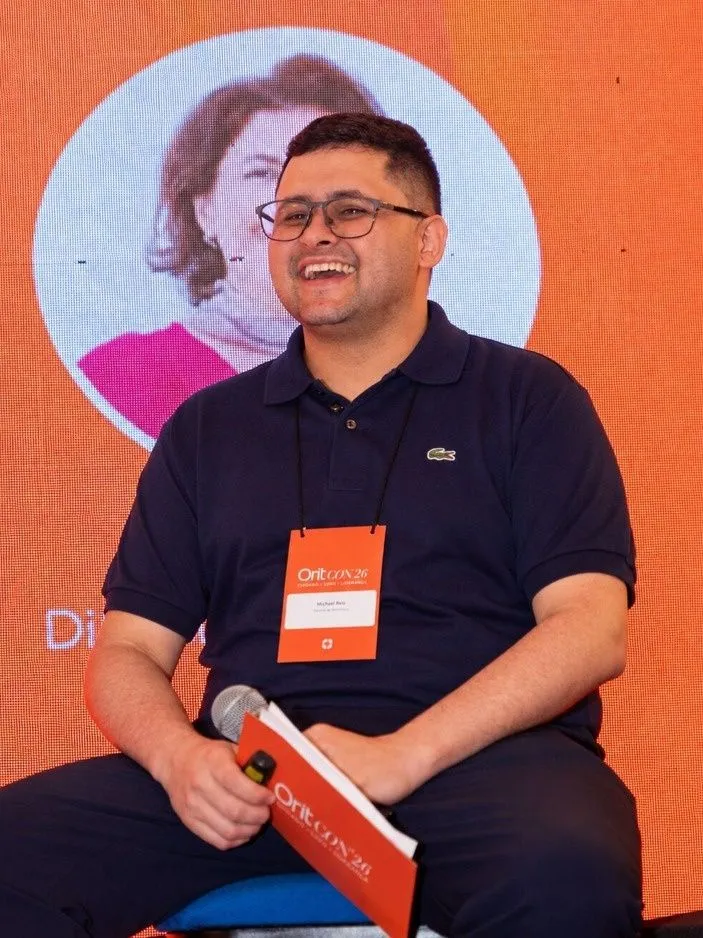 Michael Reis sorrindo no palco da OritCON'26, com o microfone e o crachá do evento